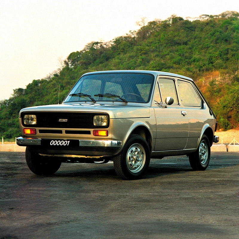 Fiat 147 na cor bege estacionado em área externa, com vegetação ao fundo. O veículo é apresentado como marco da história da Fiat no Brasil. Fiat 147 na cor bege estacionado em área externa, com vegetação ao fundo. O veículo é apresentado como marco da história da Fiat no Brasil.