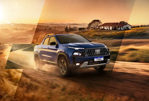 Fiat Toro azul com grade preta e logo Fiat, faróis em LED acesos, rodando em estrada de terra em cenário rural ao pôr do sol.