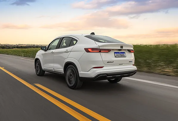 Fiat Fastback T200 Flex MHEV 12 V branco, exibido pela lateral traseira, com lanternas em LED acesas, rodando em estrada asfaltada com faixa amarela em cenário aberto.