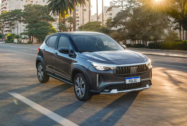 Fiat Pulse cinza com teto preto, com logo Fiat na grade dianteira e faróis em LED acesos, rodando em via urbana com prédios, árvores e luz do sol ao fundo.