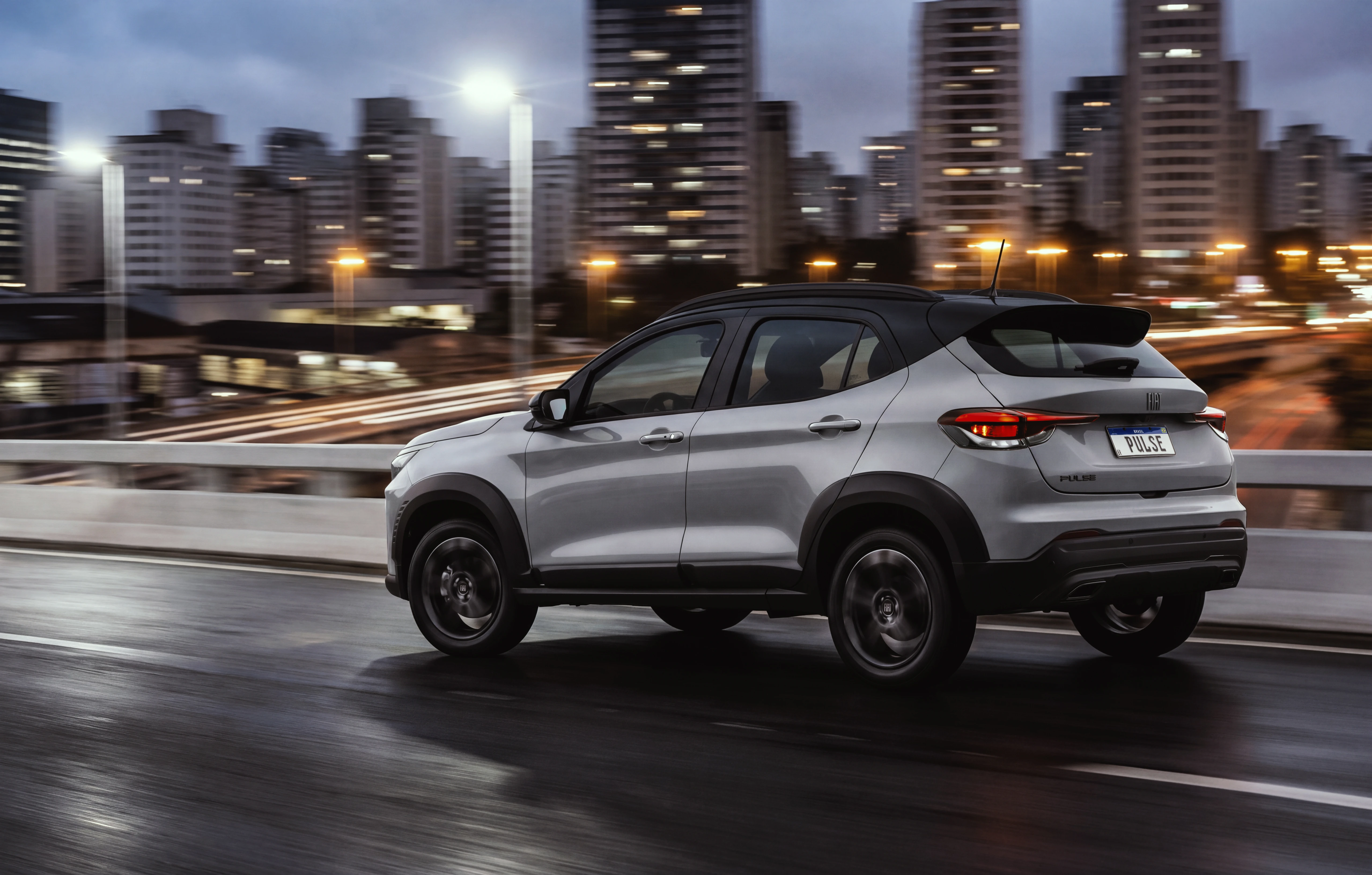 Fiat Pulse Híbrido Leve MHEV tem benefícios no uso urbano Fiat Pulse cinza com teto preto, com logo Fiat na grade dianteira e faróis em LED acesos, rodando em via urbana com prédios, árvores e luz do sol ao fundo.