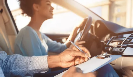 Duas pessoas com tom de pele morena dentro de um carro. Na condução, uma mulher com cabelo curto encaracolado dirige e no passageiro um homem faz anotações em uma prancheta.